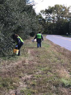 Two people in green safety vests picking up trash on the side of the road.